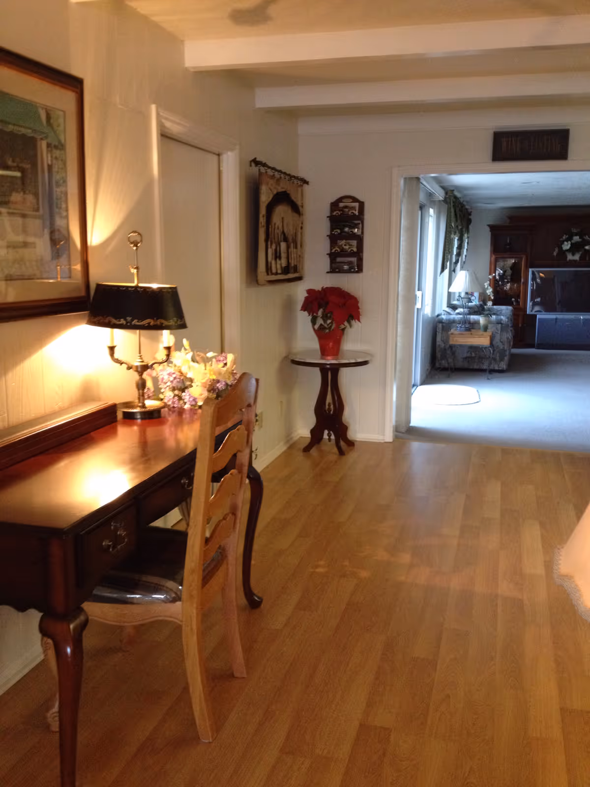 Interior hallway with a wooden desk and chair on the left, a lamp and flowers, and a small round table with a red poinsettia leading into a carpeted living room.