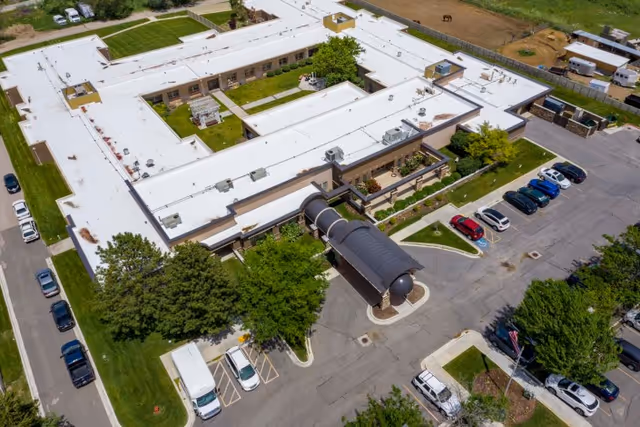 Aerial view of Copper Ridge Health Care facility showing a large, U-shaped building with a white roof, surrounded by green lawns, trees, and a parking lot with several cars parked. The entrance features a covered walkway.