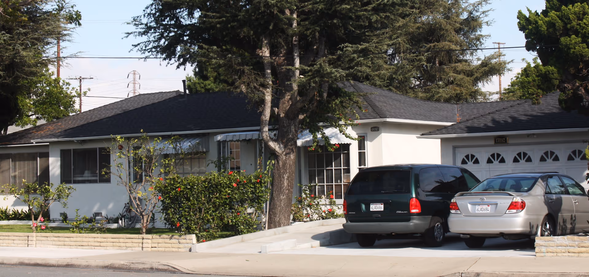 Exterior view of a single-story residential building with a dark roof, white walls, and a driveway with two parked cars. There are trees and bushes with red flowers in front of the building, and a garage with white doors is visible on the right side.