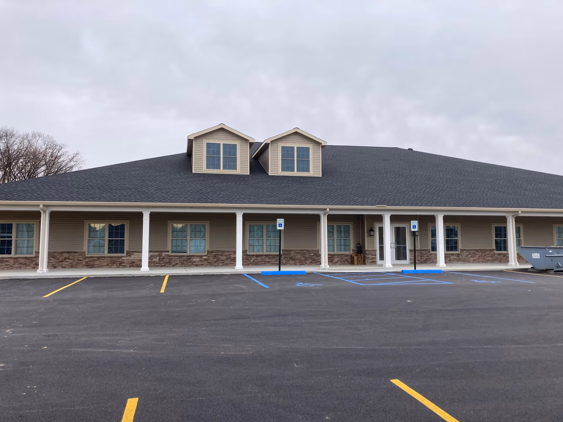 Exterior view of a single-story building with a dark gray roof and beige siding accented with stonework. The building has multiple windows with white trim and two dormer windows on the roof. There is a covered porch supported by white columns and a parking lot in front with marked handicap parking spaces. The sky is overcast.