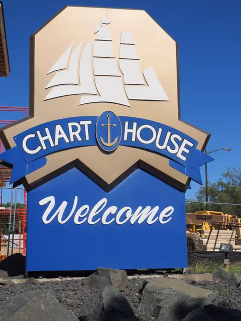 A large outdoor sign with a stylized white sailing ship at the top, the words 'CHART HOUSE' in white on a blue banner with an anchor symbol in the center, and the word 'Welcome' in cursive below on a blue background. The sign is set against a clear blue sky with some construction equipment and fencing visible in the background.