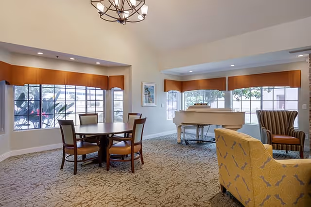 A spacious living room area with a round wooden table surrounded by four chairs, a white grand piano near large windows with brown valances, and upholstered armchairs in yellow and striped patterns. The room has beige walls, carpeted floor with a subtle pattern, and a chandelier hanging from the ceiling.
