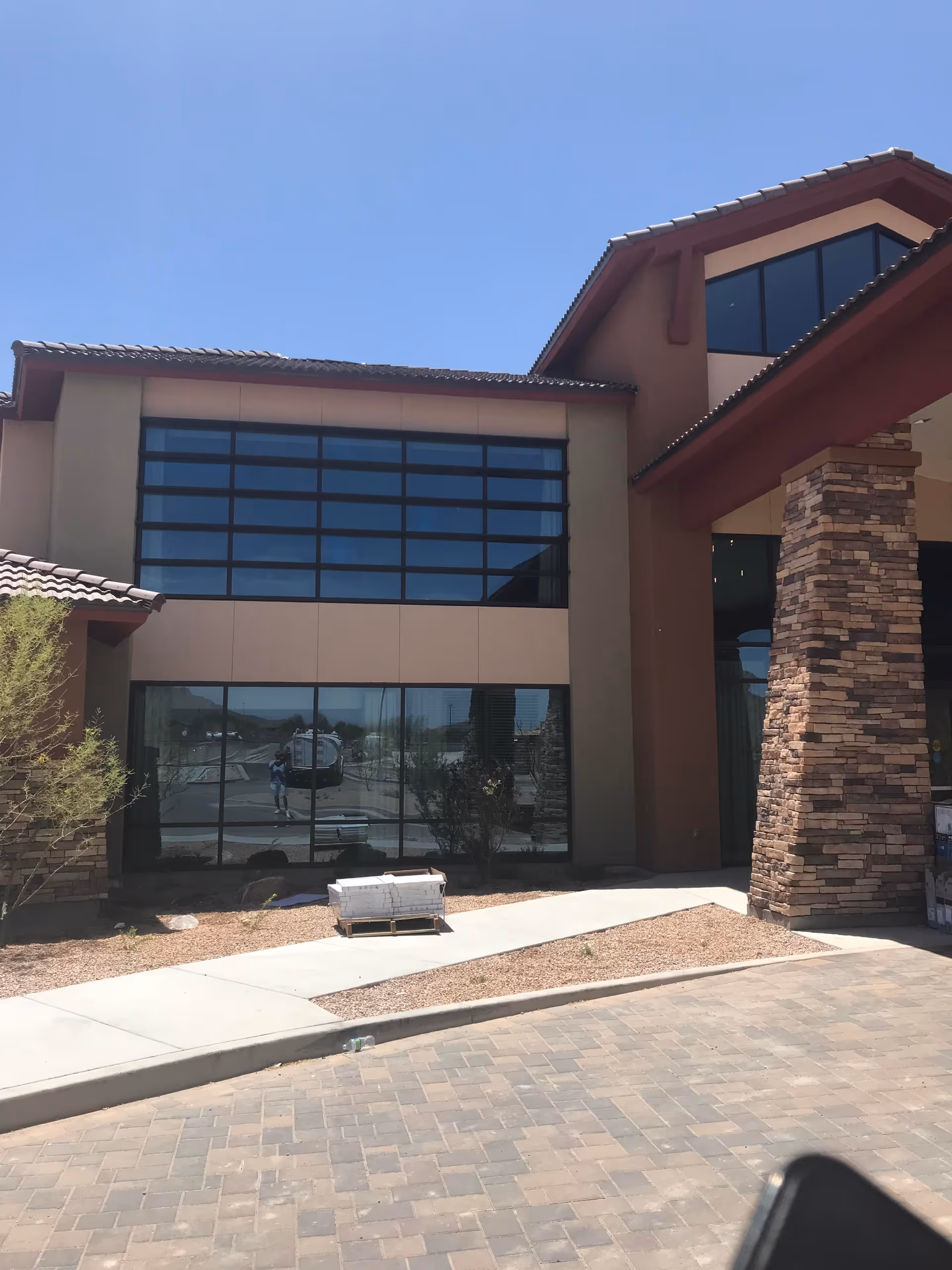 Exterior view of a building with large windows and a stone pillar at the entrance under a clear blue sky. There is a paved driveway and a sidewalk with some landscaping including small bushes and gravel.
