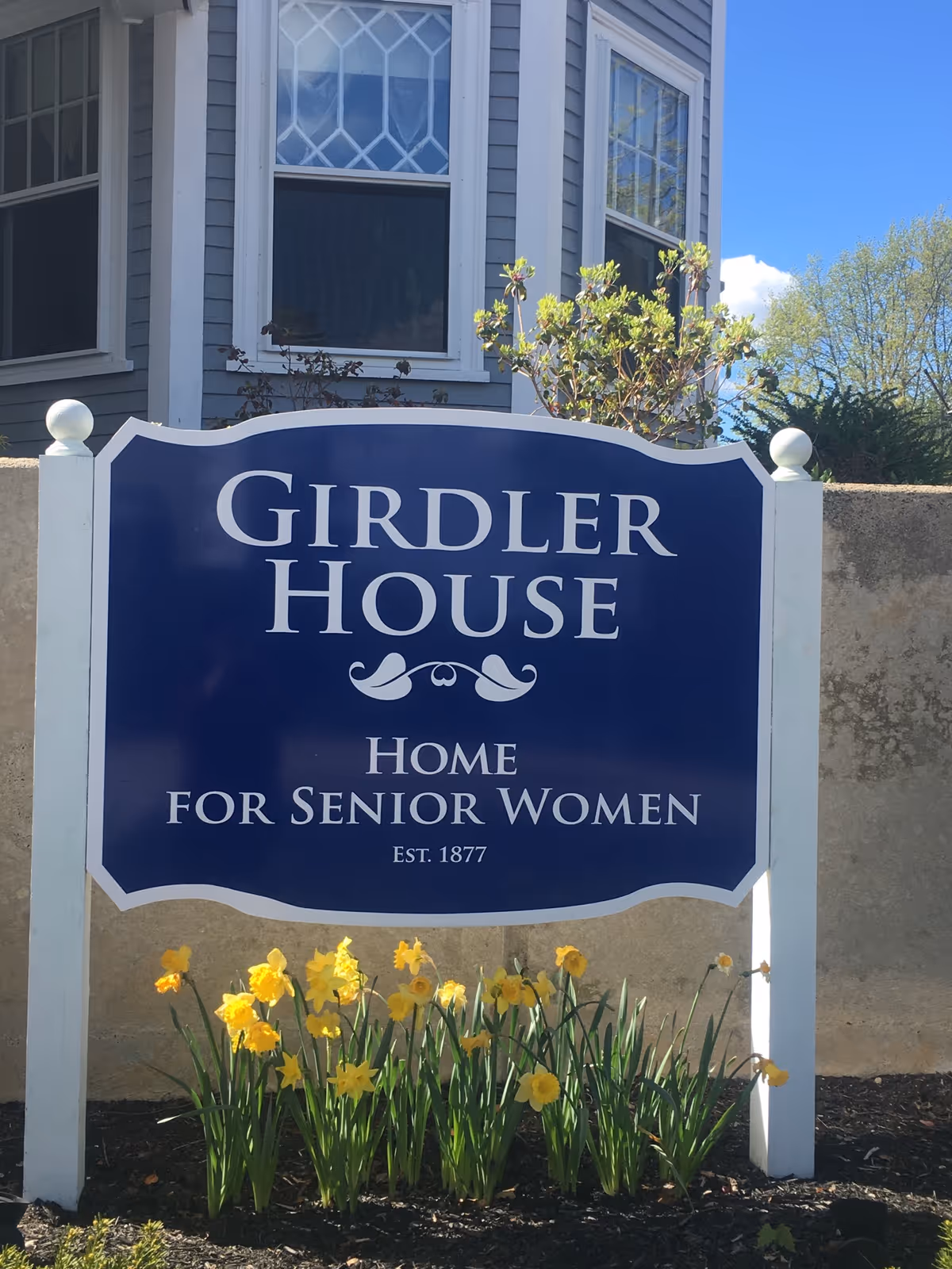 A blue and white sign in front of a building that reads 'Girdler House Home for Senior Women Est. 1877' with yellow daffodil flowers planted at the base of the sign and part of a gray building with windows in the background.