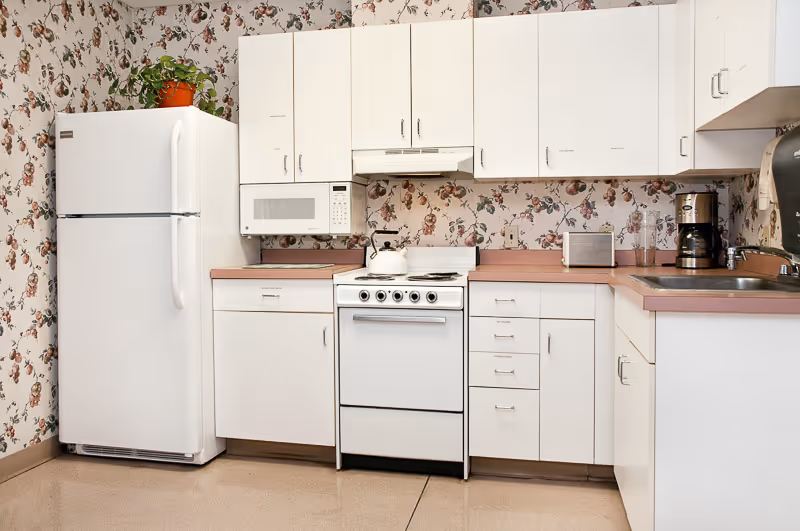 A clean kitchen with white cabinets, a white refrigerator, a white microwave, a white stove with a kettle on it, a coffee maker, a toaster, and a sink. The walls have floral wallpaper and there is a small potted plant on top of the refrigerator.