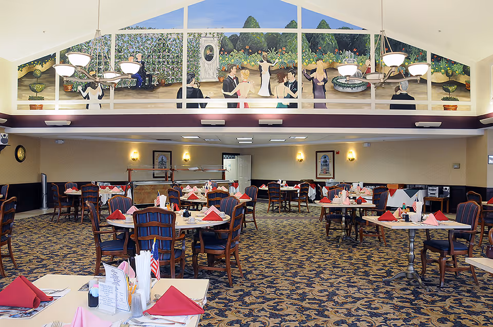 A spacious dining room with multiple tables set with red and pink napkins, utensils, and small American flags. The room has patterned carpet and upholstered chairs. On the far wall, there is a large mural depicting people dancing and socializing in a garden setting. The ceiling is vaulted with two chandeliers providing light.