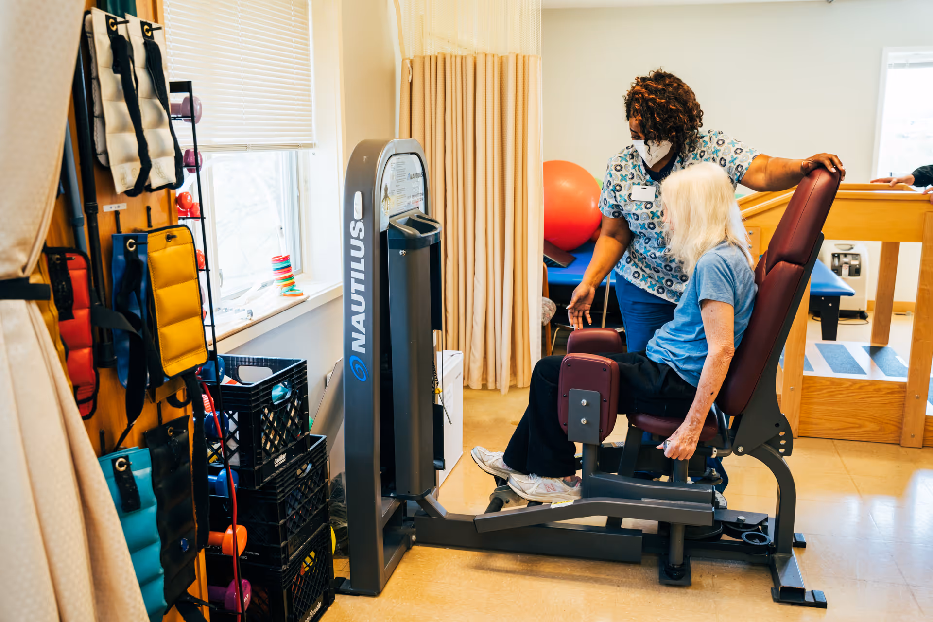 A senior woman seated on a Nautilus exercise machine in a therapy or fitness room, assisted by a healthcare worker wearing a mask and scrubs. The room contains exercise equipment, weights, and therapy balls, with a window letting in natural light.
