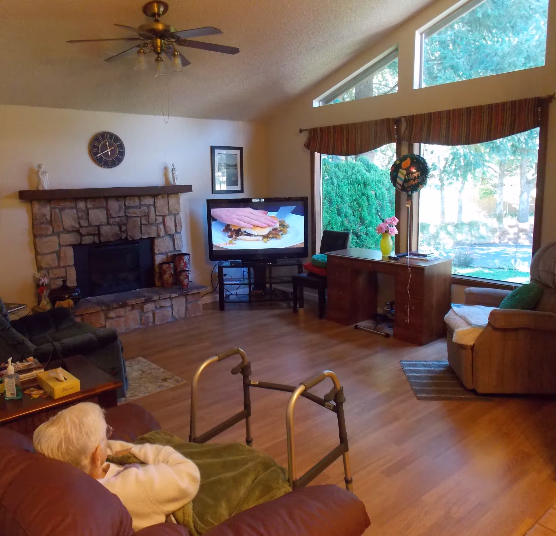 A senior living room with a stone fireplace, a ceiling fan, a television showing a cooking program, a desk with a vase of flowers, large windows with a view of greenery outside, and an elderly person sitting on a couch covered with a green blanket.