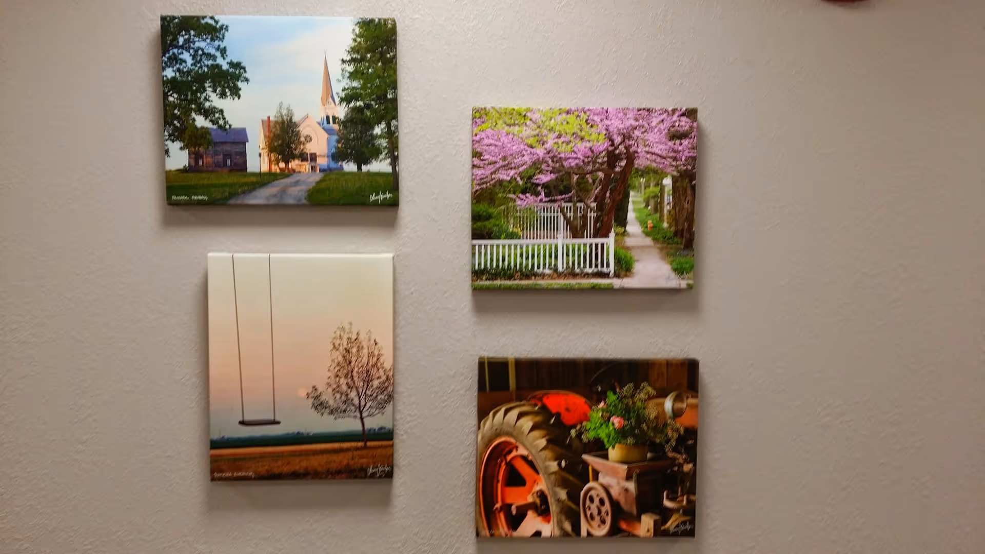 Four framed photographs arranged on a beige wall. The top left photo shows a white church with a tall steeple surrounded by trees. The top right photo depicts a pathway lined with a white fence and blooming pink trees. The bottom left photo features a swing hanging from a tree branch with a sunset or sunrise in the background. The bottom right photo shows a close-up of a vintage red tractor with a flower pot placed on it.