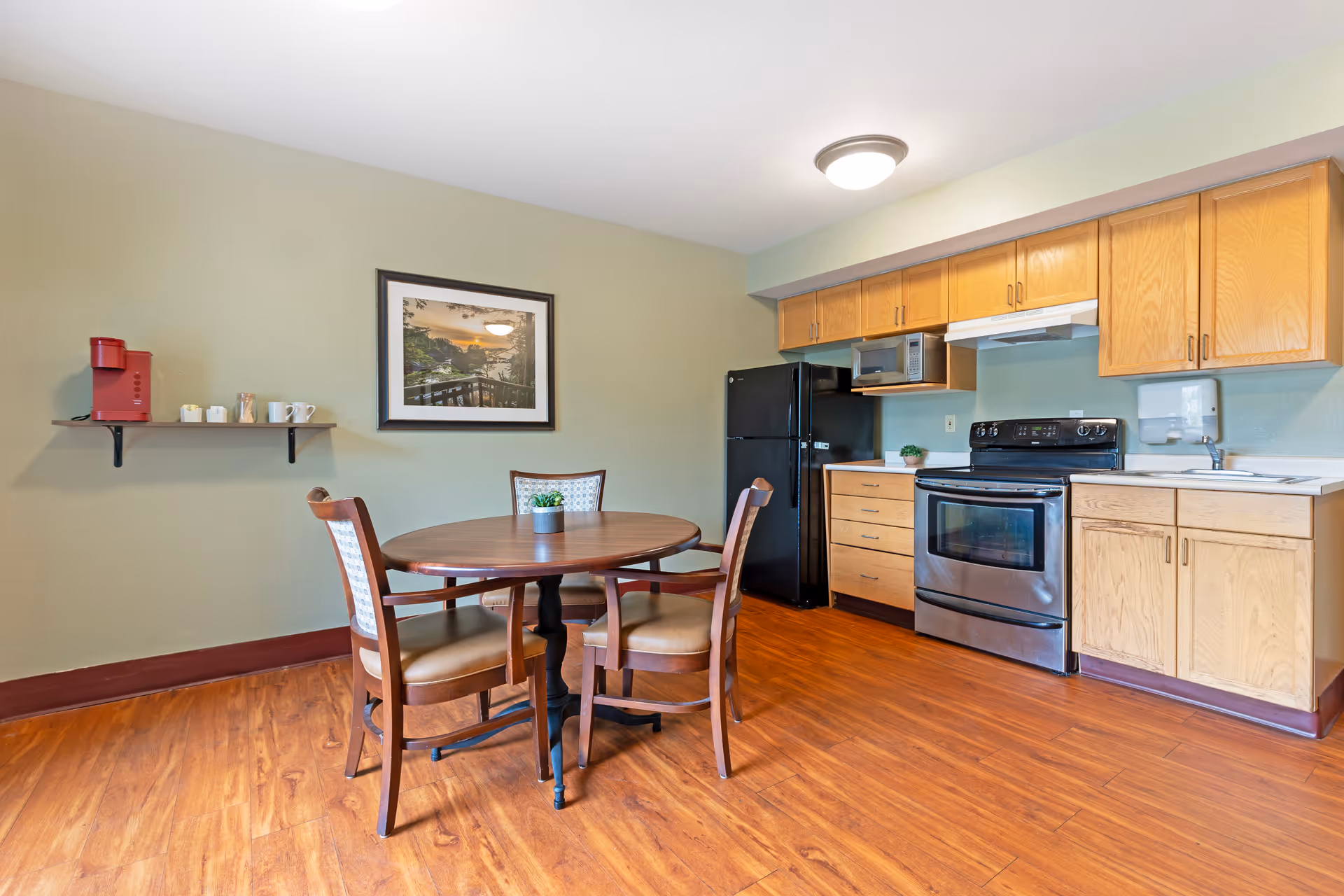 A kitchen and dining area with wooden flooring, light green walls, and wooden cabinets. The kitchen includes a black refrigerator, stainless steel oven, microwave, and a sink. A round wooden dining table with three chairs is placed in the center. A small shelf on the wall holds a red coffee maker, cups, and condiments. A framed picture hangs on the wall above the dining table.