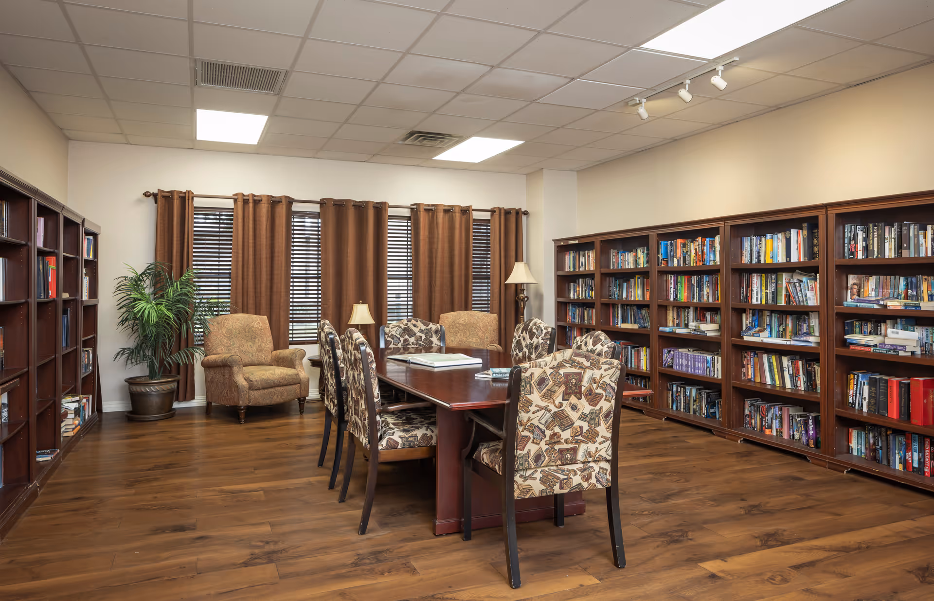 A cozy library room with wooden bookshelves filled with books lining two walls. In the center, there is a large wooden table surrounded by six upholstered chairs with patterned fabric. Two armchairs and a floor lamp are positioned near windows with brown curtains. The room has wooden flooring and a white ceiling with recessed lighting.