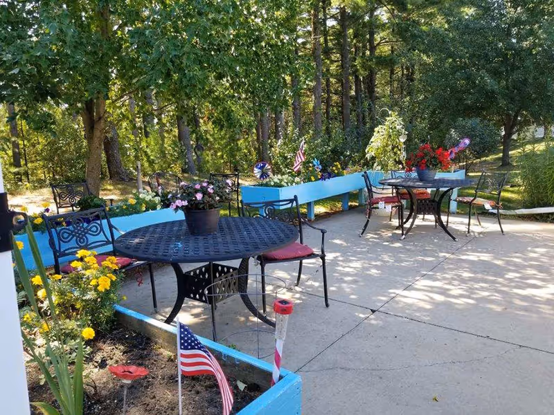 Outdoor patio area with black metal tables and chairs with red cushions, surrounded by blue planter boxes filled with colorful flowers and small American flags. Trees and greenery are visible in the background providing shade.