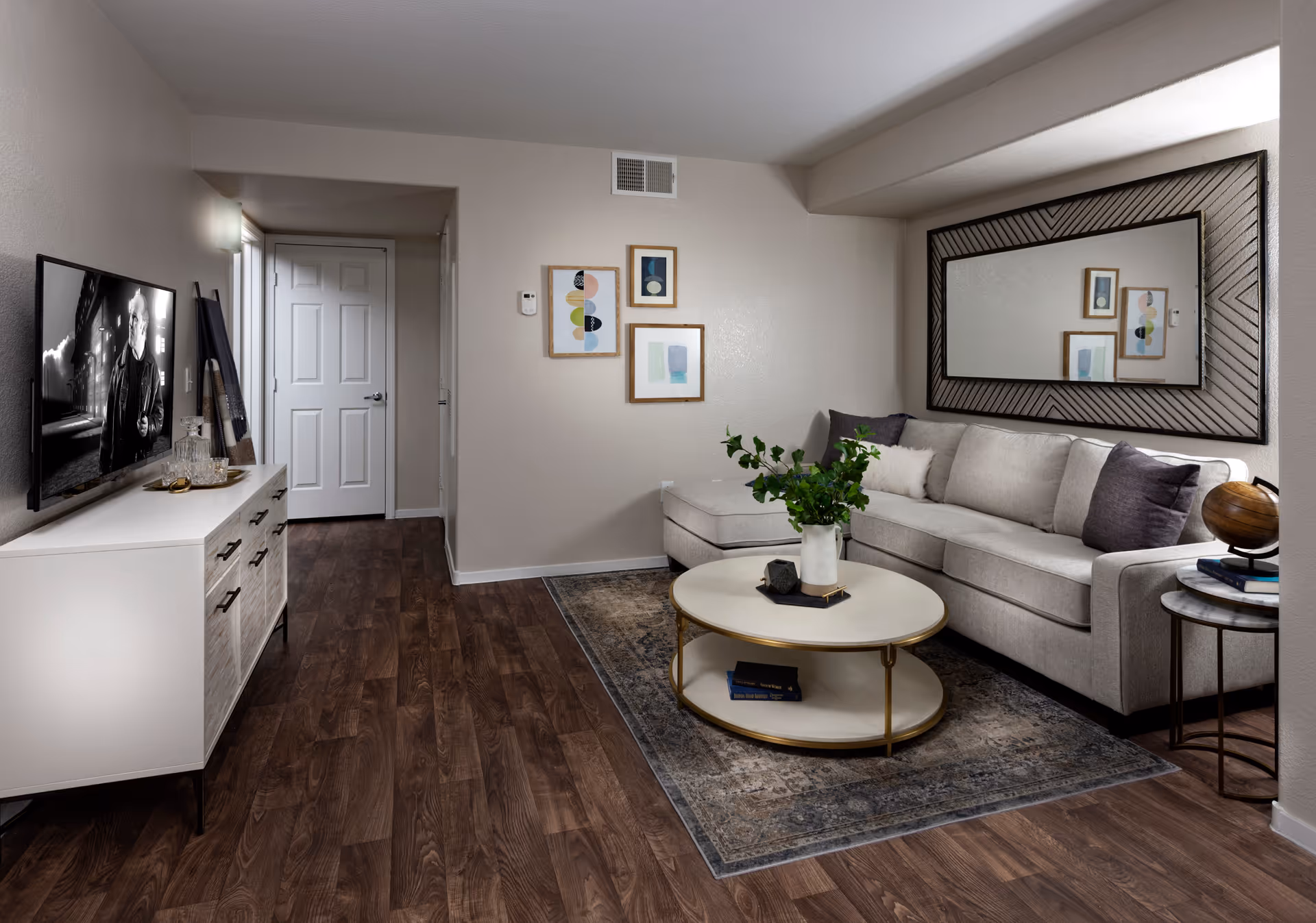 A modern living room with a light gray sectional sofa adorned with dark and light throw pillows. A large decorative mirror hangs on the wall above the sofa. In front of the sofa is a round coffee table with a white top and gold frame, holding a white vase with green foliage and a small decorative object. A patterned area rug lies underneath the coffee table. On the left side, there is a white media console with a flat-screen TV displaying a black and white image of a man. The room has wood flooring and neutral-colored walls with three framed abstract art pieces hanging on one wall. A small side table with a globe and books is next to the sofa.