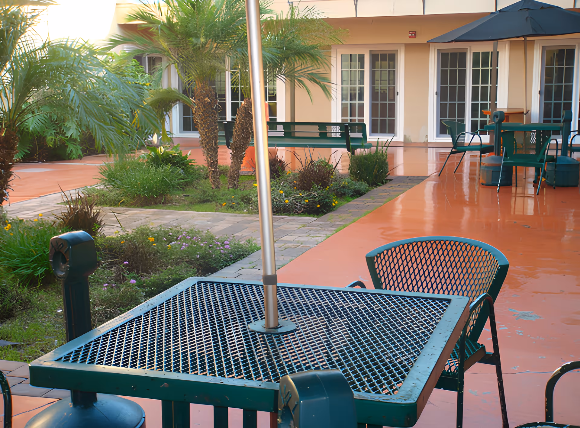 Courtyard with green metal patio tables and chairs, an umbrella pole, planted beds and a walkway in front of a building with multiple French doors.