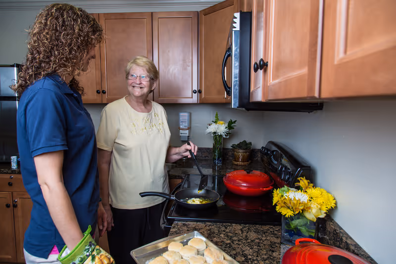 An elderly woman cooking eggs in a frying pan on a stove in a kitchen while smiling and talking to a younger woman wearing a blue shirt and oven mitts. There are biscuits on a baking tray on the counter and flowers in vases on the countertop.