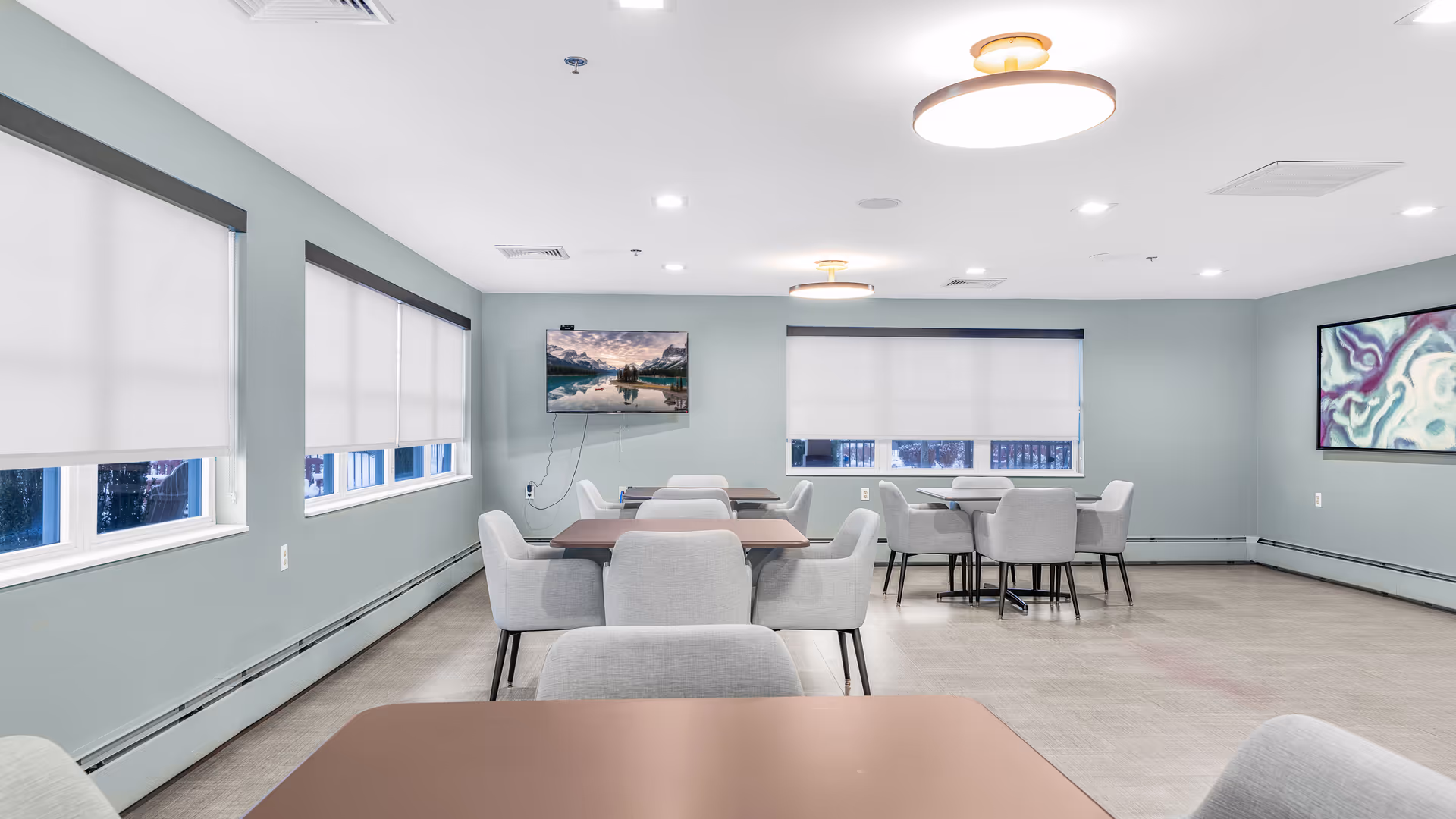 Bright communal dining/activity room with multiple tables and gray upholstered chairs, a wall-mounted TV, and large windows with blinds.