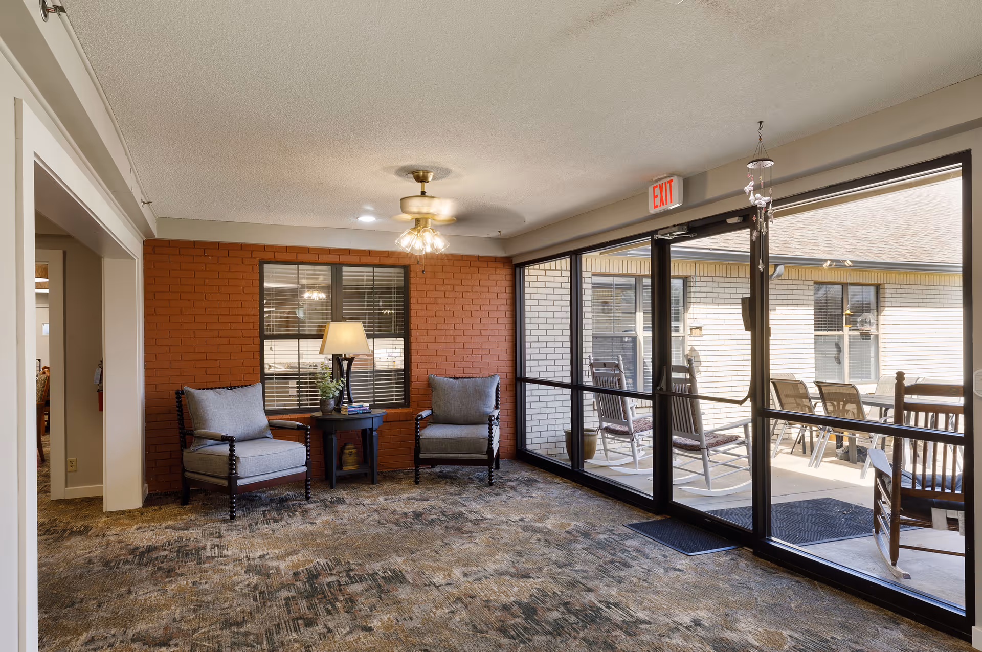A cozy sitting area inside a facility with two cushioned armchairs and a small round table with a lamp and plant between them. The room has a brick accent wall painted red, a ceiling fan with lights, and large glass sliding doors leading to an outdoor patio with rocking chairs and a table. The carpet has a patterned design, and there is an exit sign above the door.