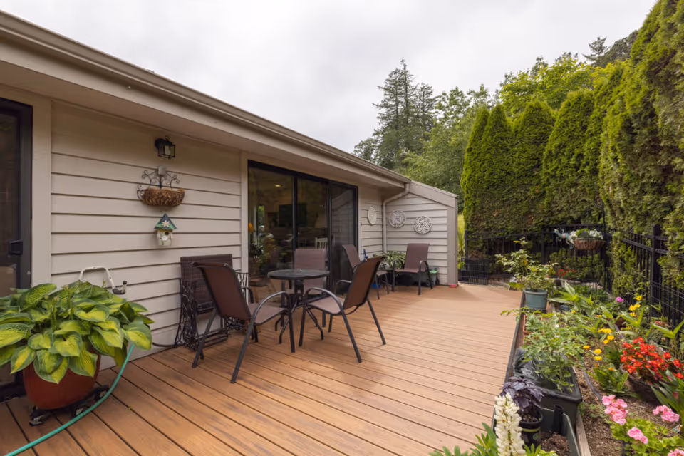 A wooden patio deck with a table and chairs, potted plants, sliding glass doors, and tall hedges along a fence.