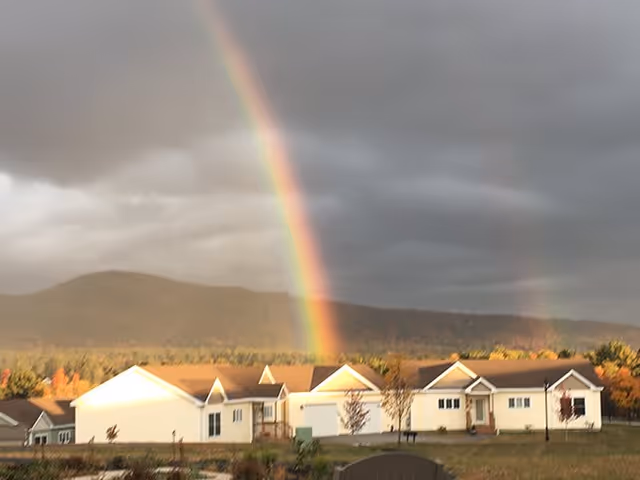 A scenic view of a residential facility with multiple single-story buildings under a cloudy sky. A vibrant rainbow arcs prominently over the buildings, with a faint second rainbow visible to the right. In the background, there are forested hills and autumn-colored trees.