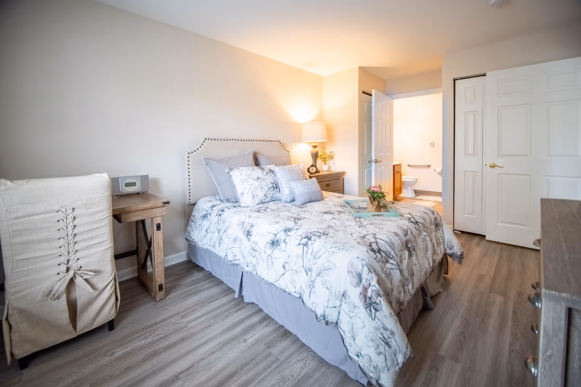 A bright bedroom with a floral-patterned bedspread on a bed with a beige upholstered headboard. There is a wooden nightstand with a lamp and decorative items next to the bed. A small wooden desk with a beige chair is positioned against the wall. The room has light-colored wood flooring and white walls. An open door reveals a bathroom with a toilet and grab bars.