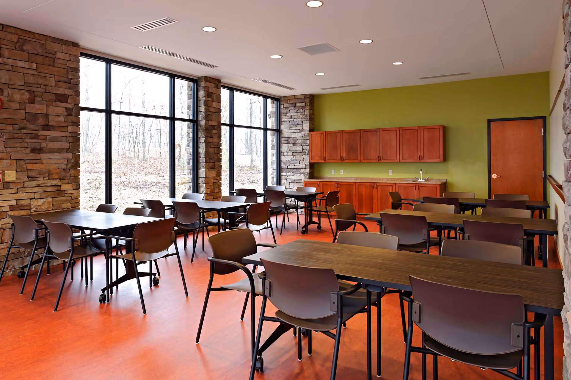 Bright communal dining room with multiple tables and chairs, large windows, stone accent walls, and cabinets along a green wall.