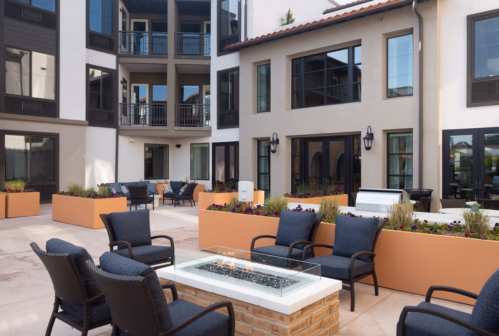 Outdoor courtyard of a senior living facility with seating around a modern glass-enclosed fire pit, planters, and surrounding apartment windows.