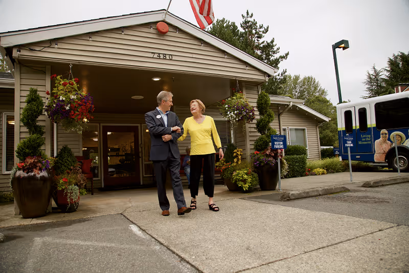 An elderly woman and a man in a suit walk arm in arm outside the entrance of a senior living facility with the address number 7480. The building has hanging flower baskets and large potted plants near the entrance. A bus with images of elderly people and the phrase 'Living in the Moment' is parked nearby. A sign indicates visitor parking.