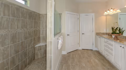 Spacious bathroom featuring a large walk-in tiled shower with a built-in bench, a double sink vanity with granite countertop, white cabinetry, a large mirror, and two closed white doors. The floor is tiled and the walls are painted a light neutral color.