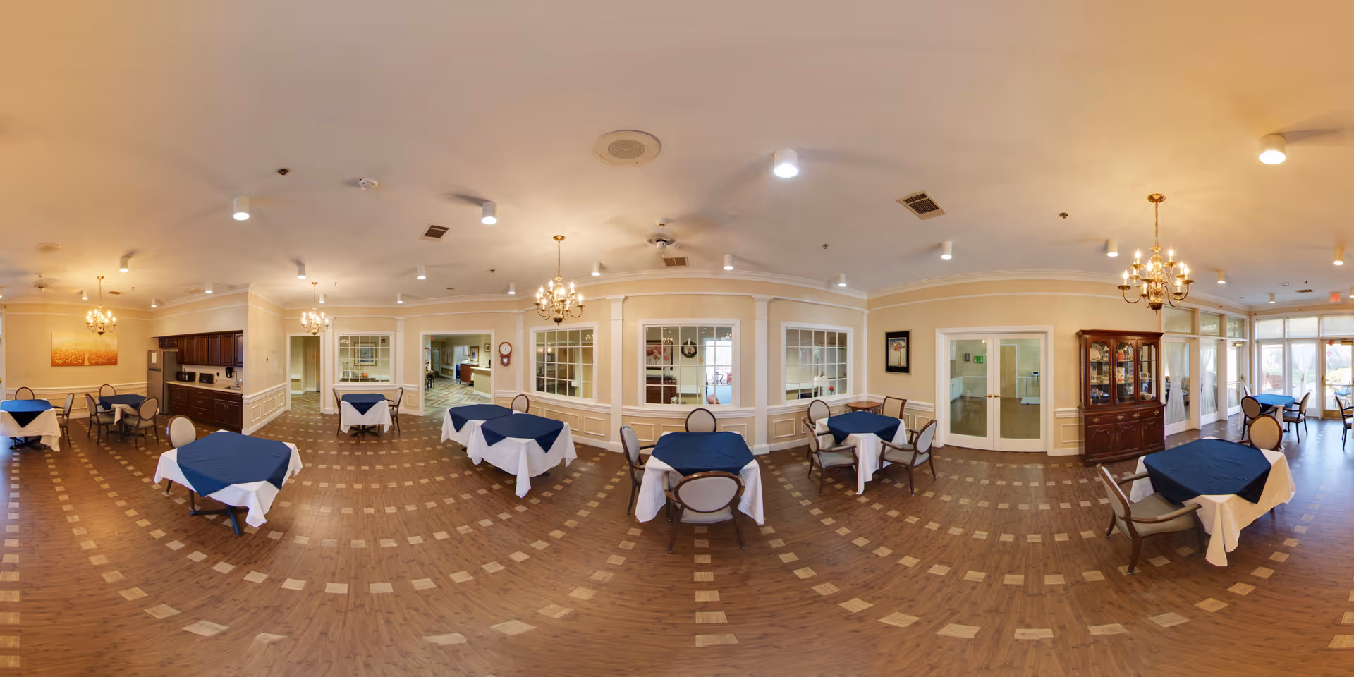 A spacious dining room with several round tables covered with white tablecloths and blue overlays. Each table is surrounded by chairs with cushioned seats and backs. The room features warm beige walls with white trim, multiple chandeliers hanging from the ceiling, and large windows letting in natural light. There is a wooden cabinet on one side and a kitchen area visible through a large serving window.