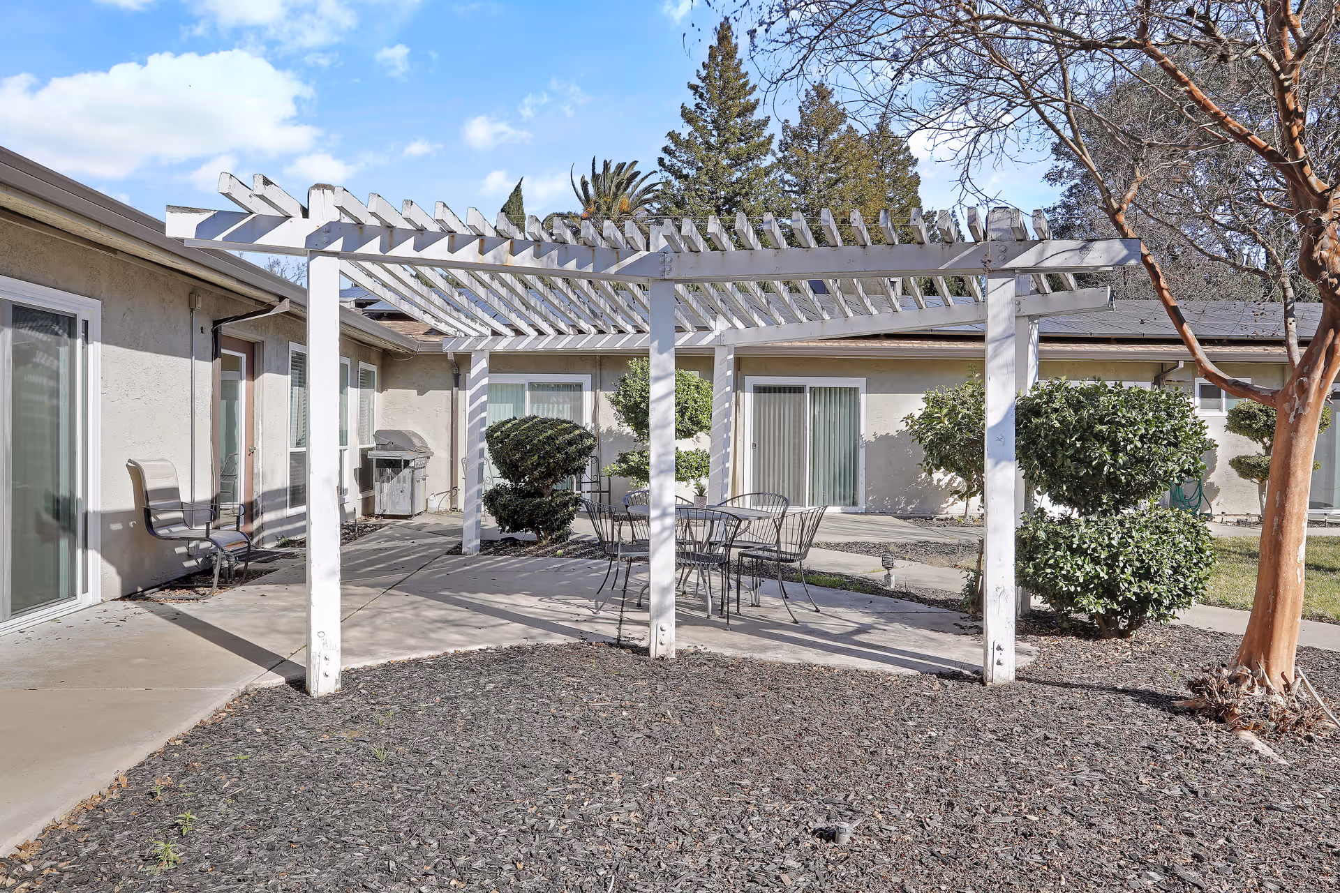 Outdoor patio area at Apple Ridge Assisted Living featuring a white wooden pergola, metal table and chairs, trimmed bushes, a tree, and sliding glass doors leading into the building under a blue sky with some clouds.