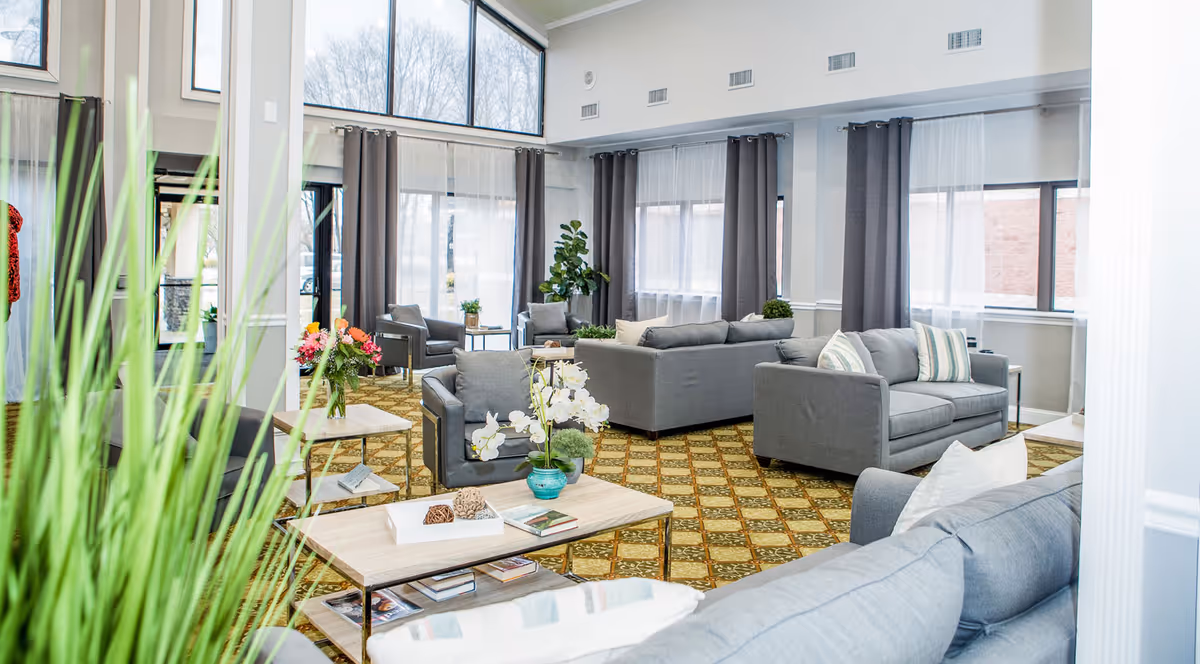 A bright and spacious living room area with multiple gray sofas and armchairs arranged around light wood coffee tables. The room features large windows with sheer white curtains and dark gray drapes, allowing natural light to fill the space. There are decorative plants and flowers on the tables and in the corners, and the floor is covered with a patterned carpet.