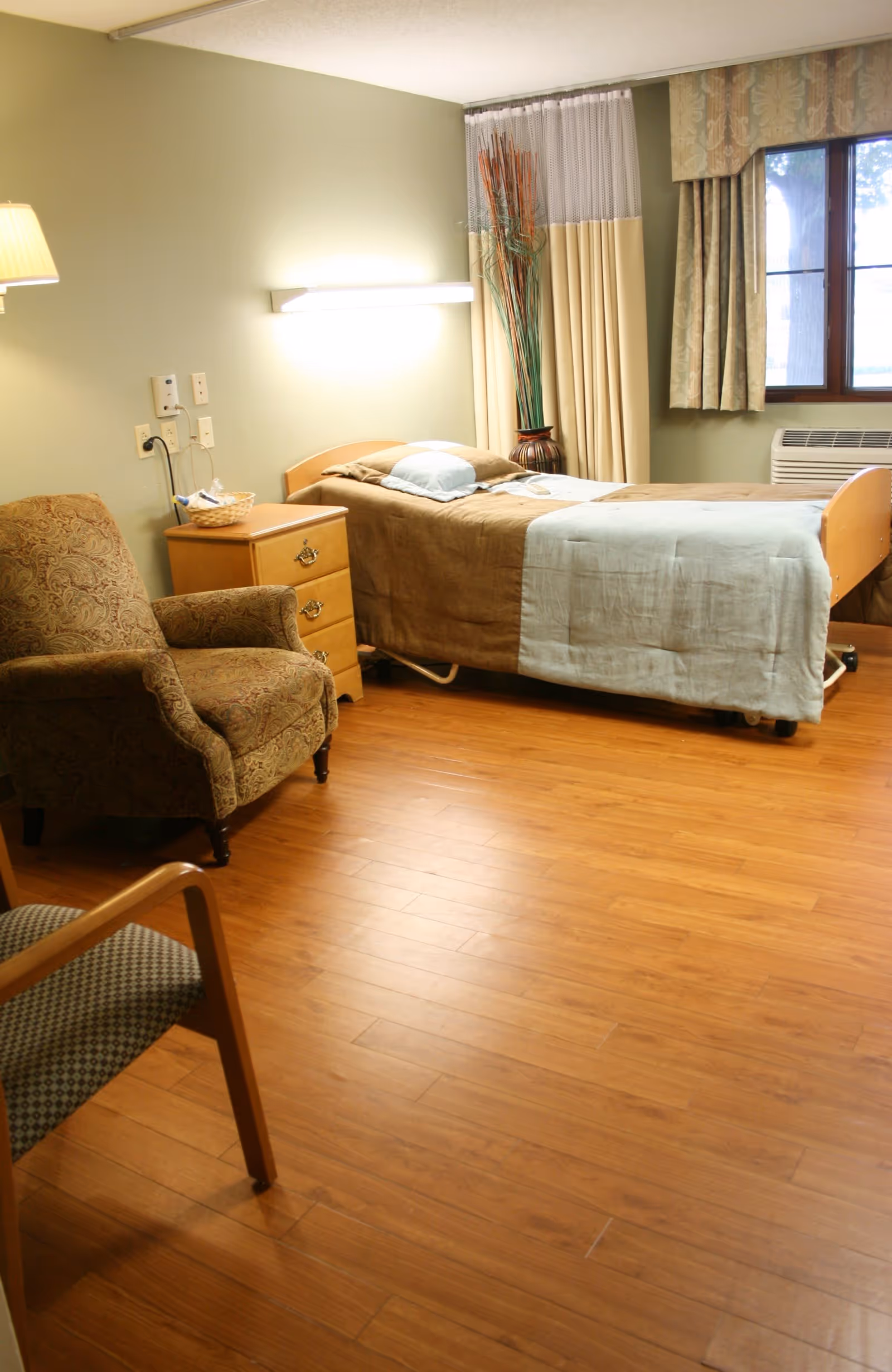 A furnished senior care bedroom with a single bed, nightstand, armchair, and hardwood floor.