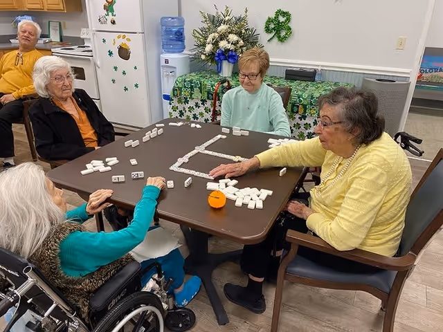 Five elderly individuals sitting around a table playing a game with dominoes in a common area decorated with St. Patrick's Day themed items including shamrocks and green decorations. The room has a kitchen area in the background with a refrigerator and a water dispenser.