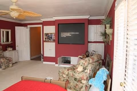 A living room with floral patterned furniture including a sofa and an armchair. The walls are painted dark red with white trim. There is a large blackboard or framed item on the wall, a ceiling fan with light, a small table with a mirror, and a window with white blinds letting in natural light.
