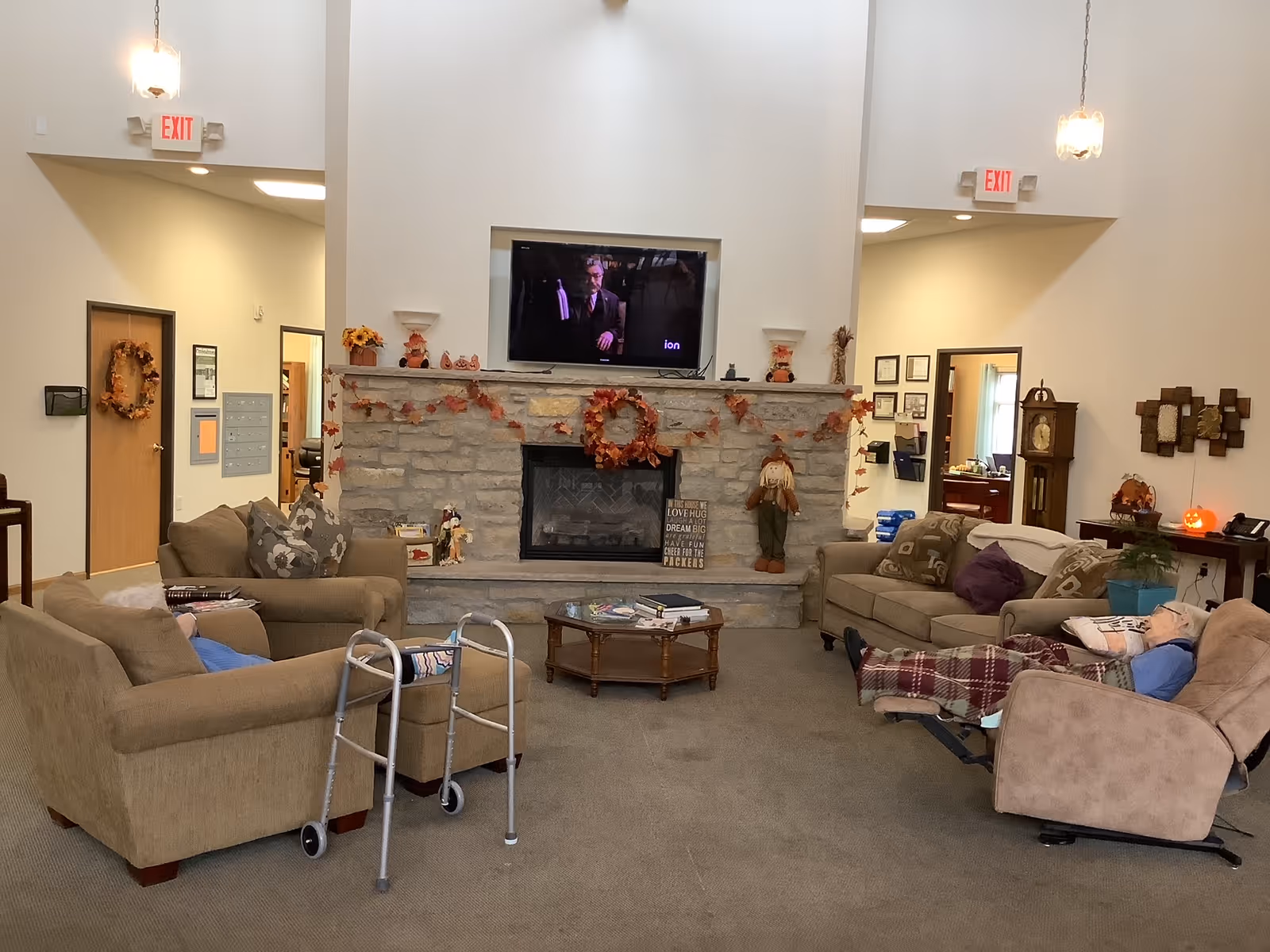 A cozy living room area in Milan Estates with beige sofas and armchairs arranged around a stone fireplace decorated with autumn-themed garlands and a wreath. A flat-screen TV is mounted above the fireplace. There is a walker in front of one armchair, and an elderly person is reclining in a chair with a blanket. The room has high ceilings with hanging lights and exit signs above doorways leading to other rooms.
