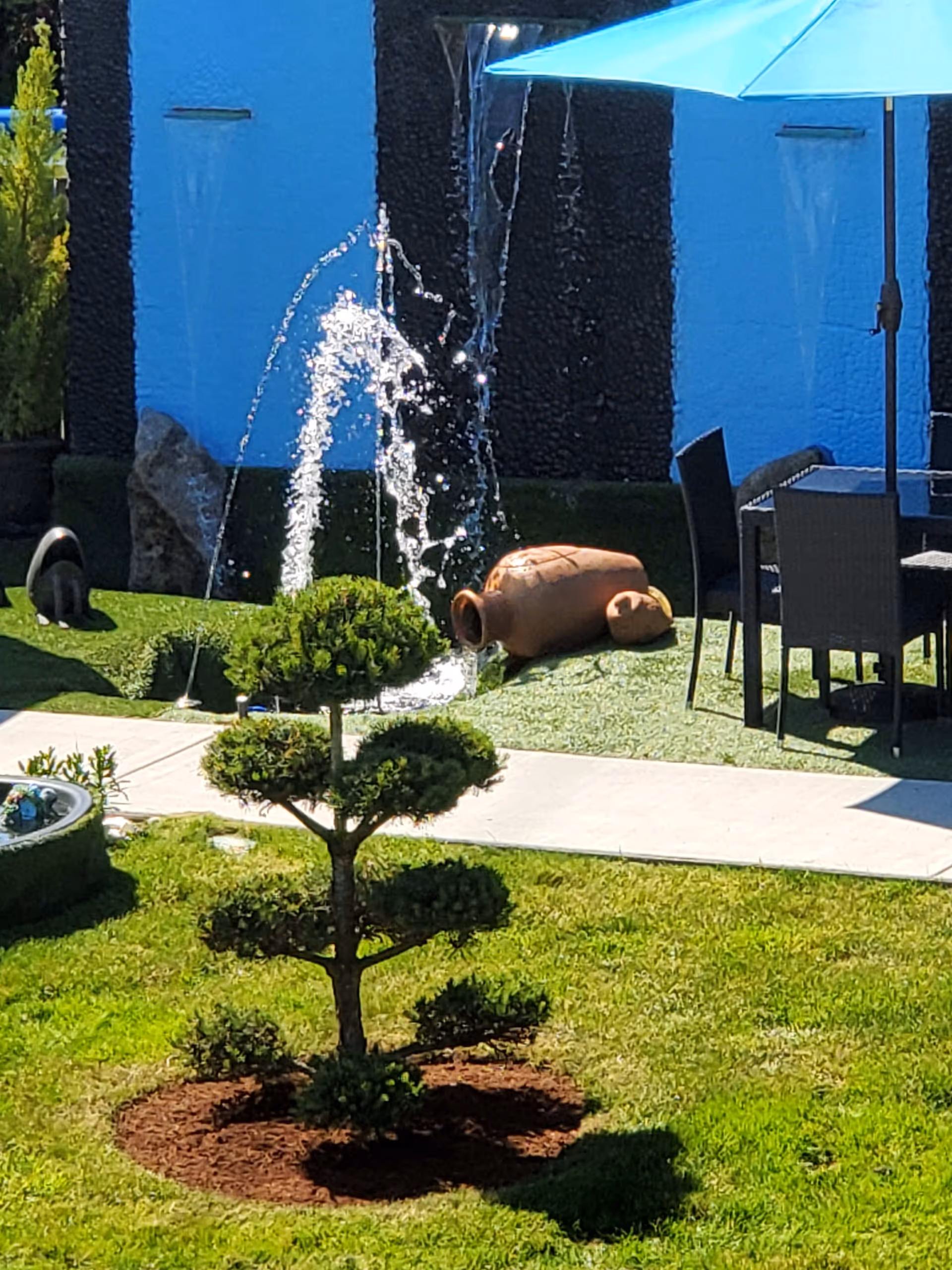 A well-maintained outdoor garden area with a small, neatly trimmed tree in the foreground. Behind the tree, there is a water fountain with water jets creating arcs of water. To the right, there is a patio area with a table, chairs, and a large blue umbrella providing shade. The background features a blue and black textured wall with water flowing from spouts.