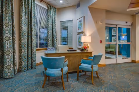 A cozy reception area with a wooden desk and two blue upholstered chairs. Behind the desk is a window with patterned curtains and blinds. A table lamp and a small flower arrangement sit on the desk. The area has carpeted flooring with a blue and beige pattern, and there is a glass door entrance visible in the background.