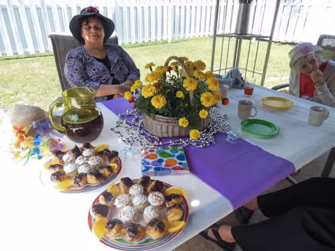 Two elderly women sitting at a table outdoors under a canopy. The table is covered with a white and purple tablecloth and decorated with a basket of yellow flowers, plates of pastries, a pitcher of dark beverage, and several cups. A white picket fence and green grass are visible in the background.