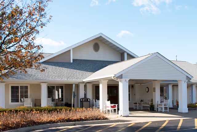 Exterior view of a single-story senior living facility building with a covered entrance, white siding, and several white rocking chairs on the porch. There are some bushes and a tree with autumn leaves in front of the building under a clear blue sky.