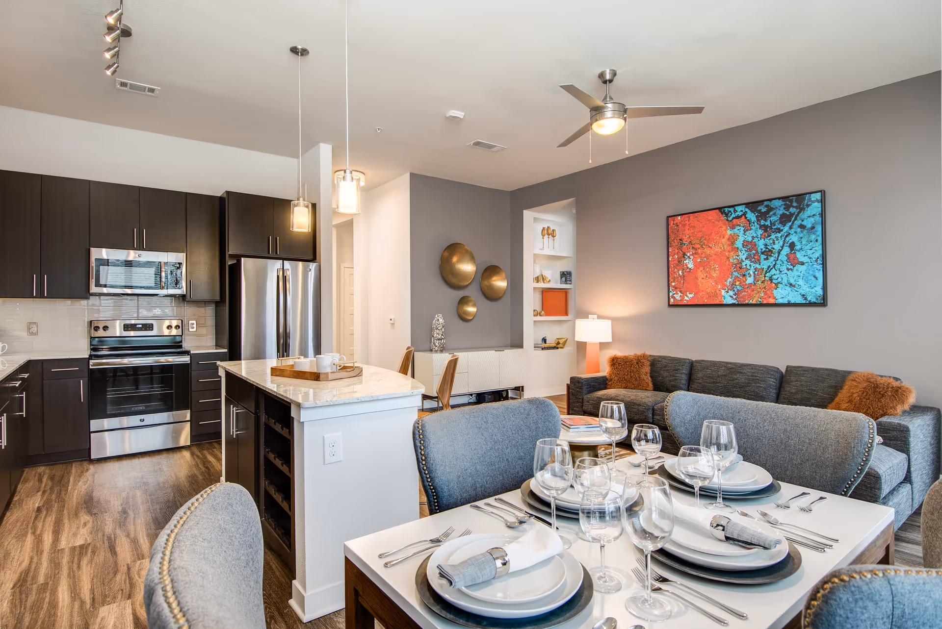 Modern open-concept living space featuring a kitchen with dark cabinets, stainless steel appliances, and a marble island. In the foreground, a dining table is set with plates, glasses, and silverware. The living area includes a gray sofa with orange pillows, a ceiling fan, and colorful abstract wall art.
