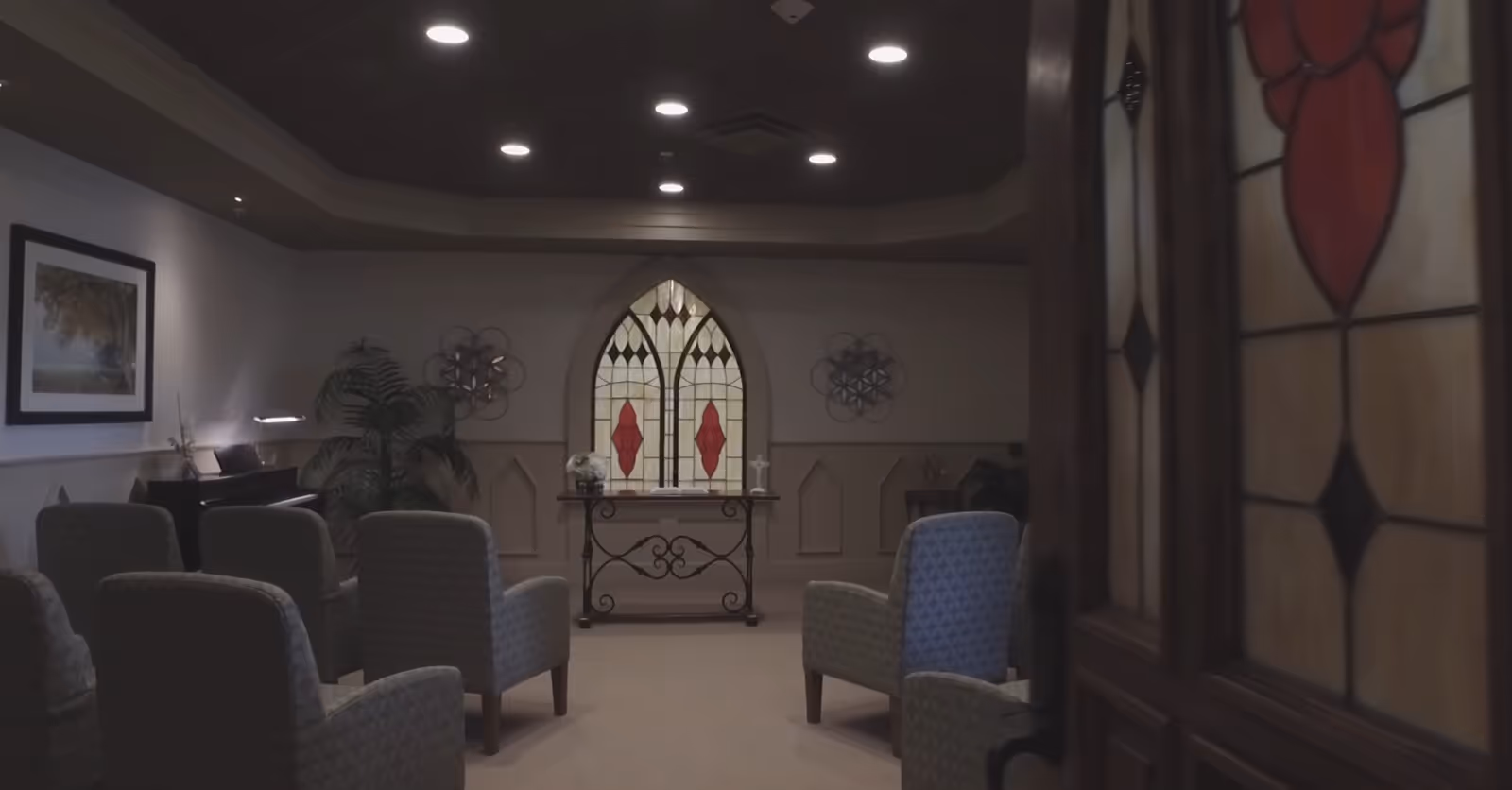 A small chapel-like interior with upholstered chairs arranged facing a stained-glass window and a small altar table.