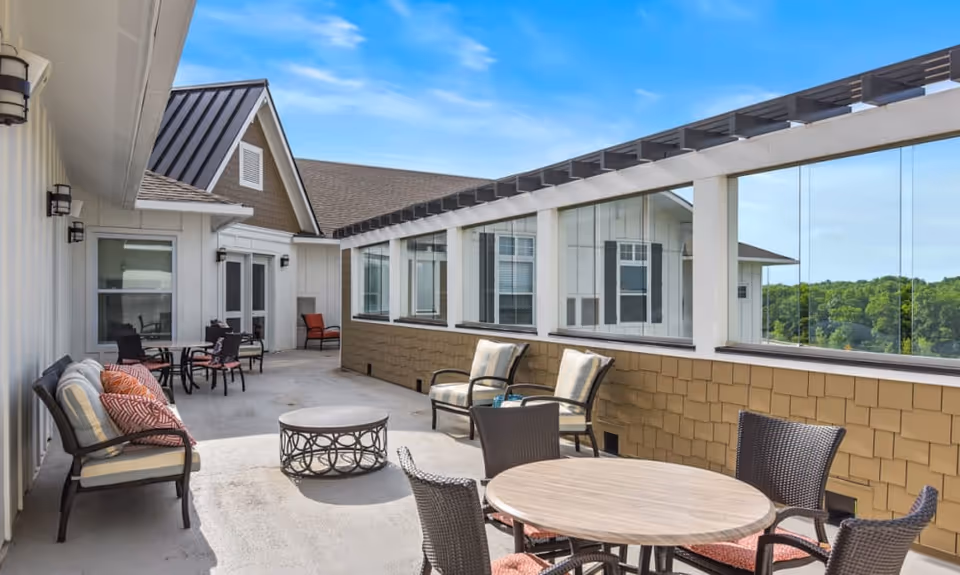 Outdoor rooftop patio with multiple seating areas, tables, and glass windbreaks along a senior living building overlooking trees.