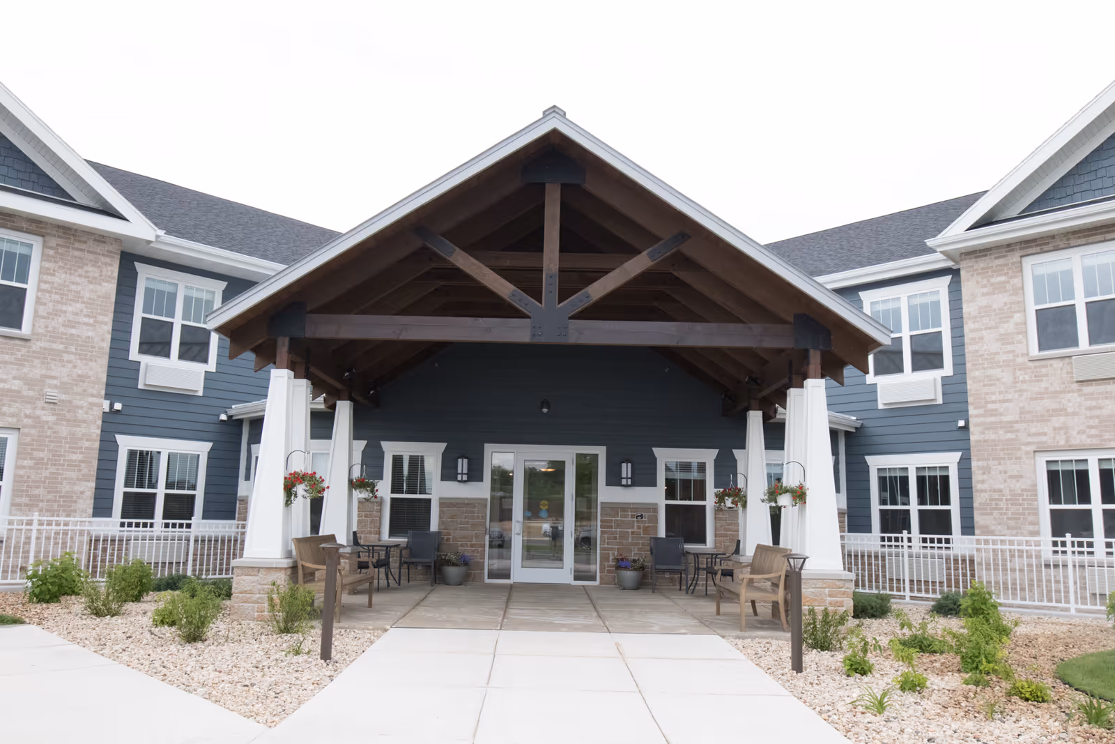 Entrance of a senior living facility with a covered porch supported by white columns and dark wooden beams. There are benches and chairs with small tables on the porch, hanging flower baskets, and landscaped areas with small plants and rocks on either side of the walkway leading to glass double doors.