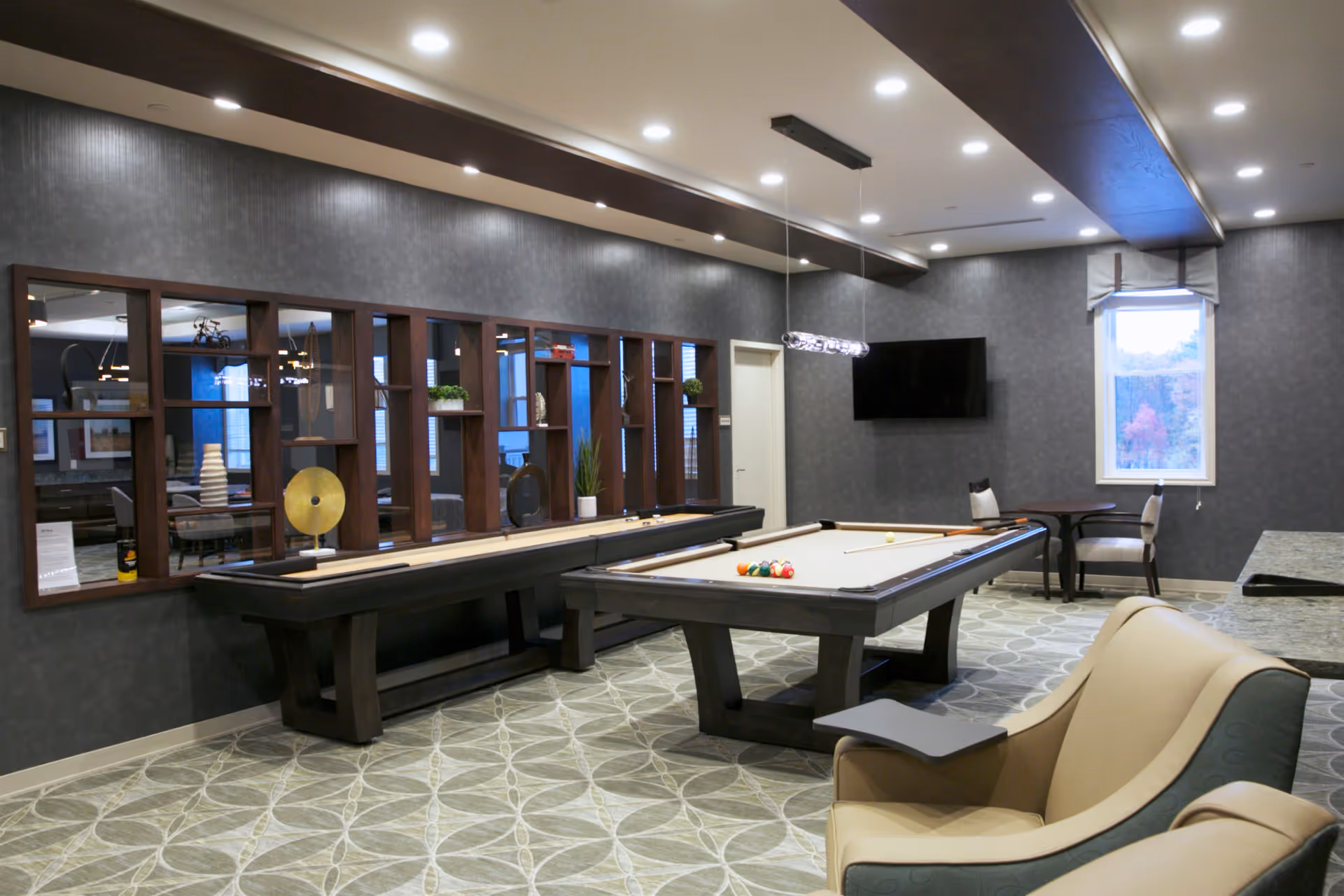 A modern game room featuring a pool table with balls and cues, a shuffleboard table along the wall with decorative items on a shelf above it, a wall-mounted flat-screen TV, a small table with two chairs near a window, and a comfortable armchair in the foreground. The room has patterned carpet, recessed ceiling lights, and dark accent beams on the ceiling.