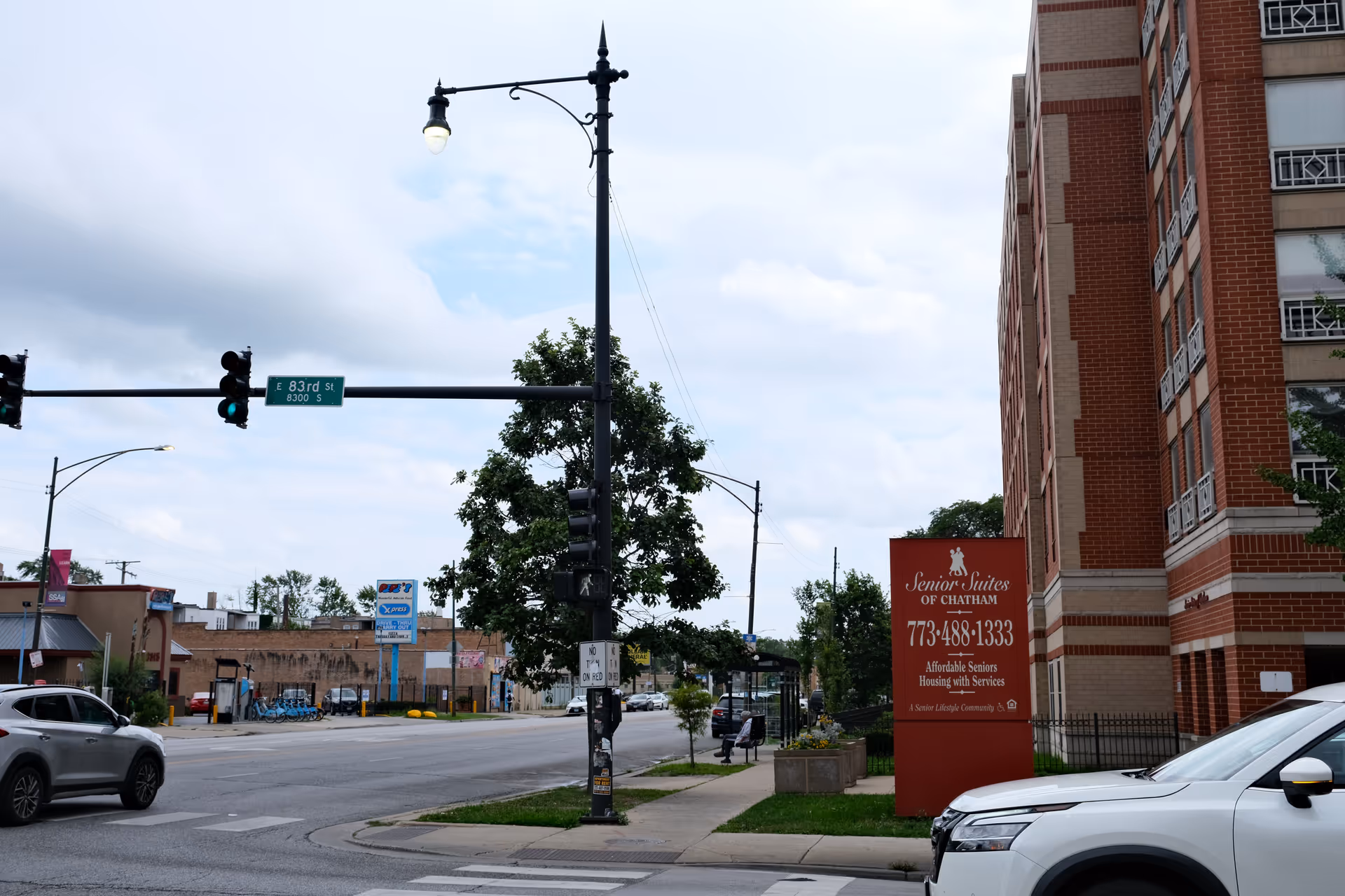 Street view showing a traffic light and street sign for E 83rd St with a red sign for Senior Suites of Chatham next to a multi-story brick building. The sign displays the phone number 773-488-1333 and advertises affordable seniors housing with services. Cars are parked along the street and a person is sitting at a bus stop in the background.