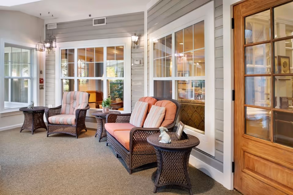 A cozy indoor seating area with wicker furniture including a cushioned armchair and loveseat with striped pillows, two wicker side tables with decorative items, large windows with white frames, and a wooden door with glass panes.
