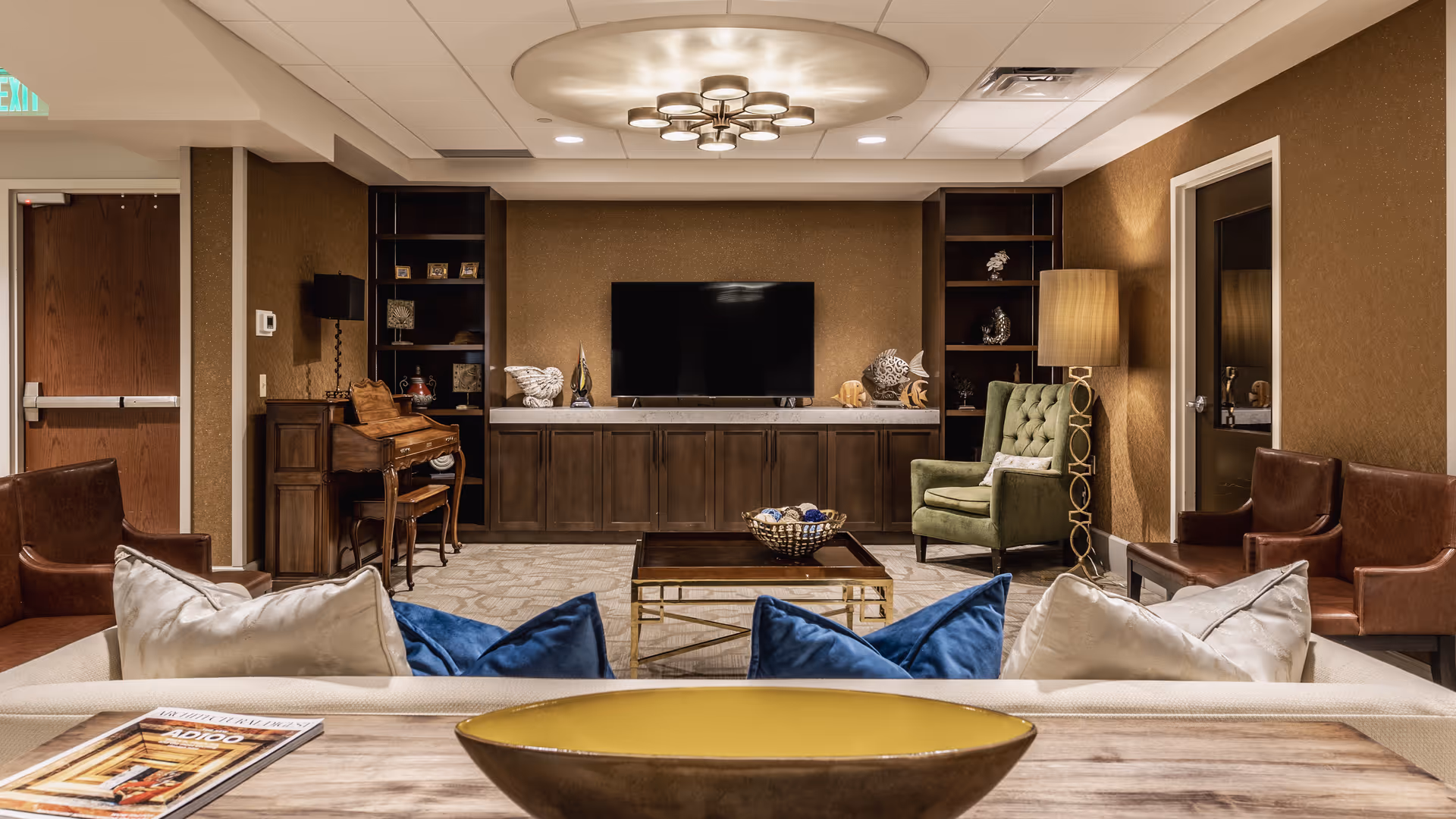 A cozy living room with a beige sofa adorned with blue and white pillows facing a flat-screen TV mounted on a brown wall. The room features a wooden coffee table with a decorative bowl, a green armchair with a pillow, two brown leather chairs, a wooden desk with a chair, and built-in shelves with decorative items. A modern ceiling light fixture illuminates the space.