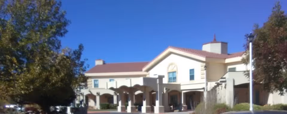 Front exterior of a two-story senior living building with a covered entrance, arched columns, red tile roof and surrounding trees under a clear blue sky.