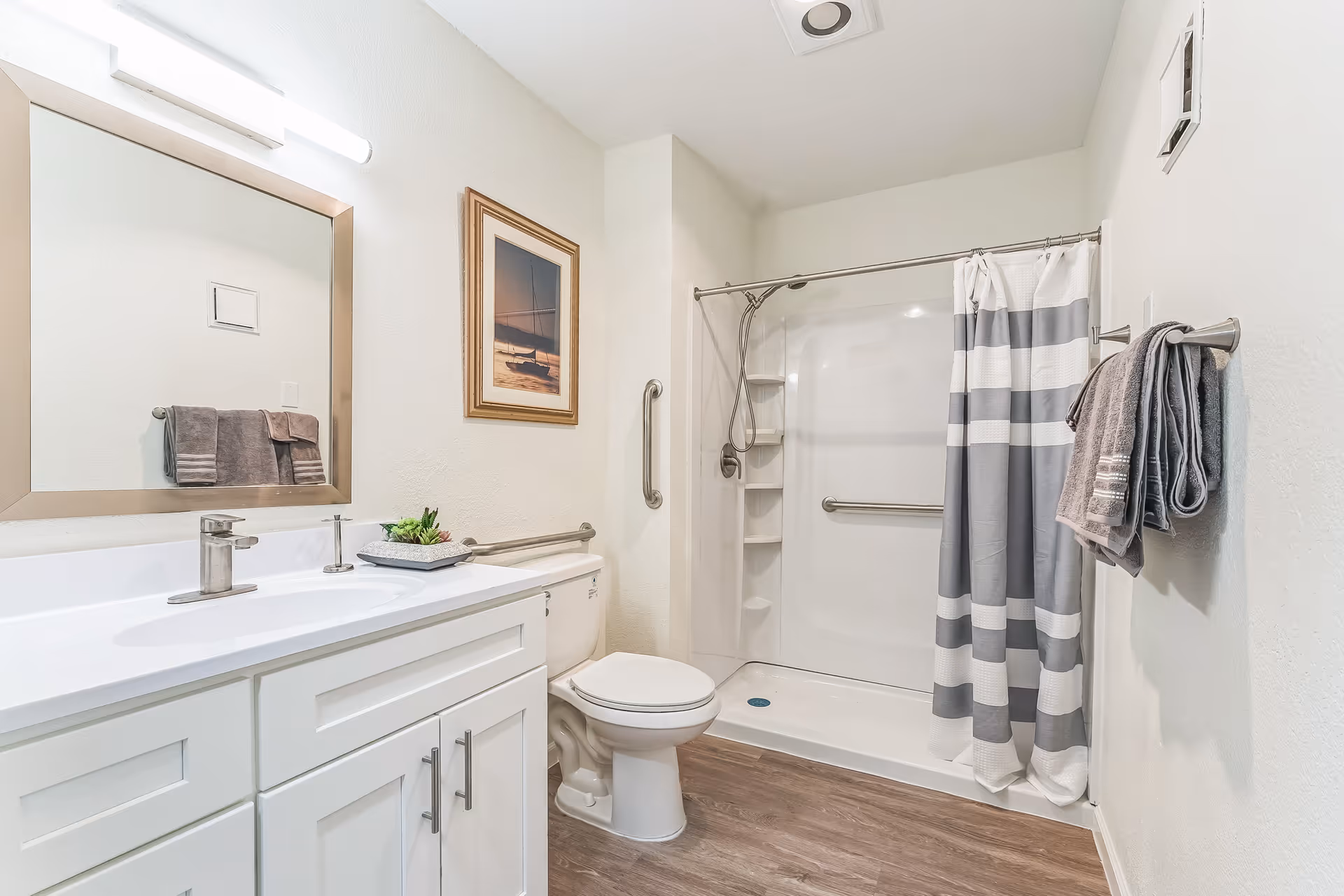 Bright and clean bathroom with a white vanity and sink, a large mirror above the sink, a toilet, and a walk-in shower with a gray and white striped shower curtain. There are gray towels hanging on a towel rack and a framed picture on the wall. The floor has wood-like flooring.