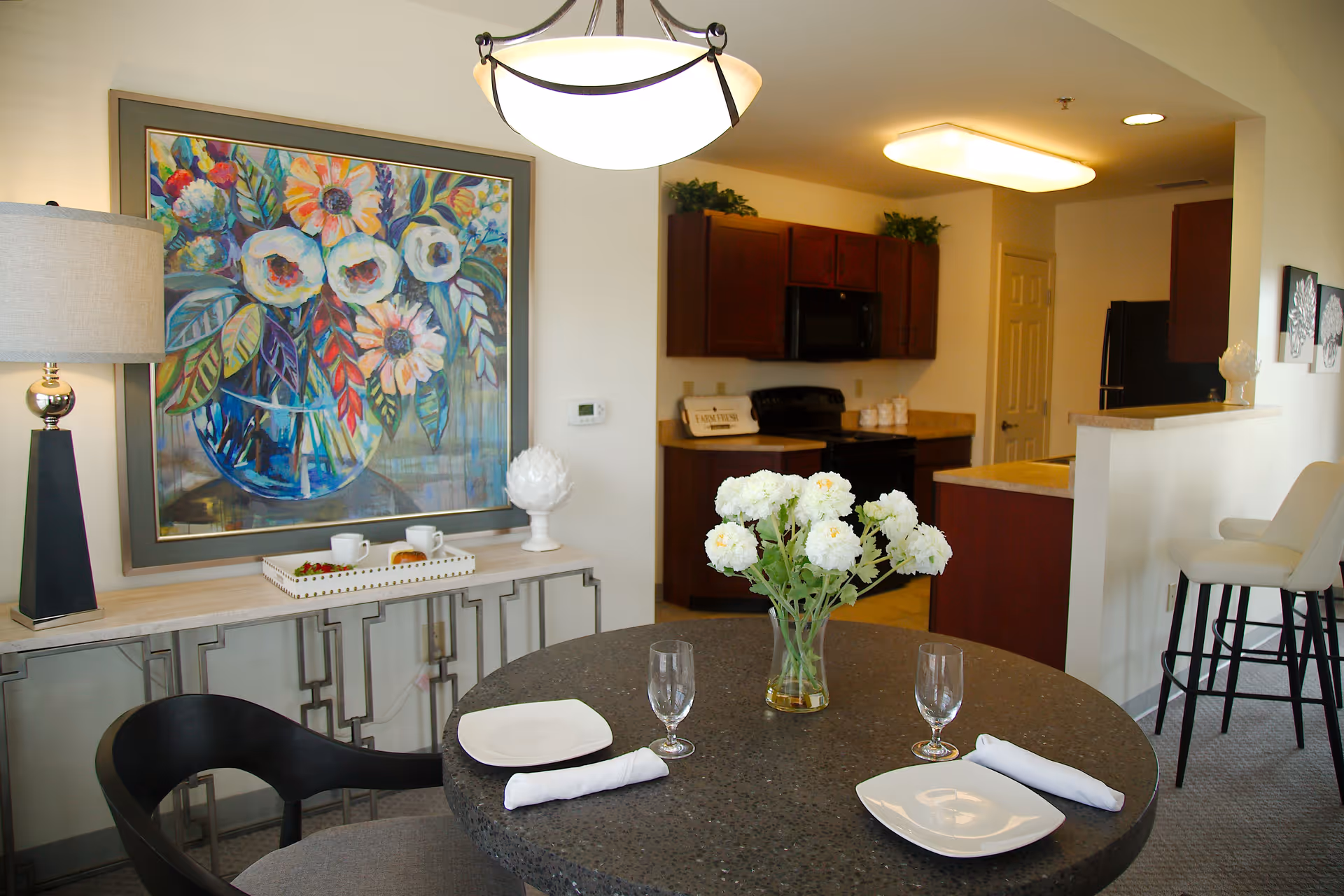 A dining area with a round table set for two with white plates, napkins, and glasses. A vase with white flowers is in the center of the table. Behind the table is a console table with a lamp, a tray with cups and food, and a large colorful floral painting on the wall. In the background, there is a kitchen with dark wood cabinets, a stove, microwave, and refrigerator. Two white bar stools are positioned at a counter separating the kitchen and dining area.